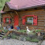 Drewniany dom z bali z czerwonymi okiennicami, otoczony kwiatami w donicach, rustykalnymi dekoracjami i zielenią ogrodu.