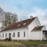 Ponadstuletni biały dom na Mazurach z ceglanym dachem, tradycyjnymi oknami ze szprosami i starymi drzewami wokół.