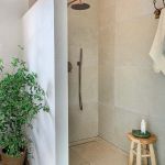Minimalistyczny prysznic walk-in z kamiennymi płytami, miedzianą armaturą, drewnianym stołkiem i rośliną w koszu.