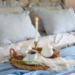 Wiklinowa taca z filiżanką kawy, białą porcelaną, konwaliami i zapaloną świecą na łóżku z błękitnym pledem.