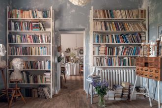 Eklektyczne wnętrze domowej biblioteki z wysokimi regałami pełnymi książek, surowymi szaroniebieskimi ścianami i drewnianą podłogą. Charakter przestrzeni budują rzeźbiarskie popiersia, stary katalog szufladkowy, świece, stosy książek oraz bukiet polnych k