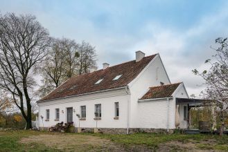 Ponadstuletni biały dom na Mazurach z ceglanym dachem, tradycyjnymi oknami ze szprosami i starymi drzewami wokół.