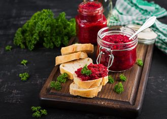 Dwa otwarte słoiki z tartymi buraczkami oraz kromki jasnego chleba z buraczkami i pietruszką na drewnianej desce.