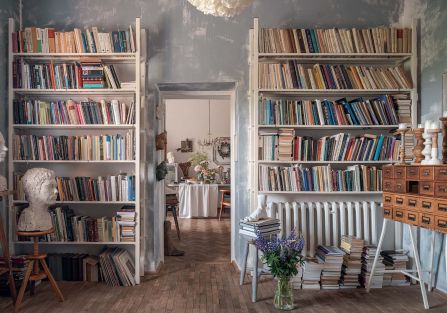 Eklektyczne wnętrze domowej biblioteki z wysokimi regałami pełnymi książek, surowymi szaroniebieskimi ścianami i drewnianą podłogą. Charakter przestrzeni budują rzeźbiarskie popiersia, stary katalog szufladkowy, świece, stosy książek oraz bukiet polnych k