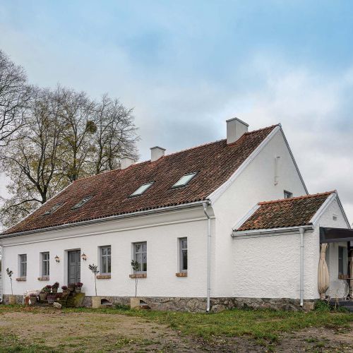 Ponadstuletni biały dom na Mazurach z ceglanym dachem, tradycyjnymi oknami ze szprosami i starymi drzewami wokół.