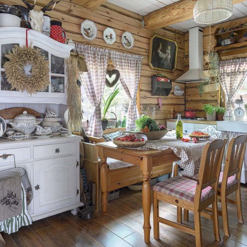 Rustykalna kuchnia w drewnianym domu z białymi meblami, stołem z litego drewna, zasłonami w kratę i dekoracjami w stylu country.