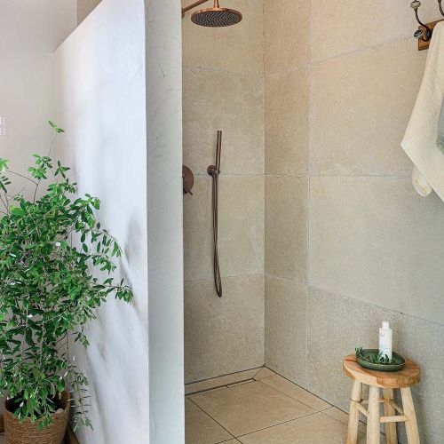 Minimalistyczny prysznic walk-in z kamiennymi płytami, miedzianą armaturą, drewnianym stołkiem i rośliną w koszu.