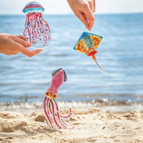 Kolorowe szklane dekoracje w kształcie meduzy, płaszczki i kałamarnicy na plaży.