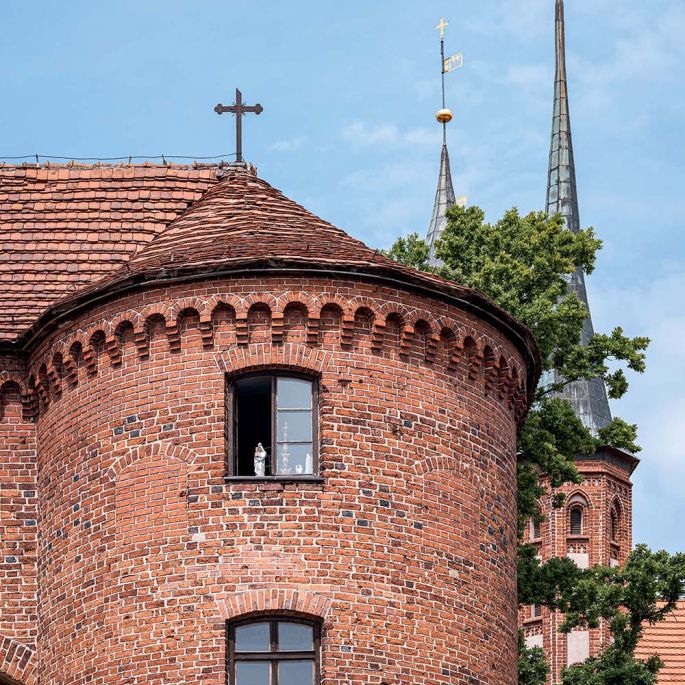 Eklektyczne wnętrze domowej biblioteki z wysokimi regałami pełnymi książek, surowymi szaroniebieskimi ścianami i drewnianą podłogą. Charakter przestrzeni budują rzeźbiarskie popiersia, stary katalog szufladkowy, świece, stosy książek oraz bukiet polnych k