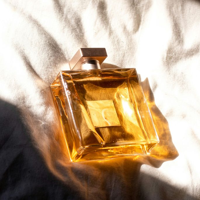 Elegancki, przezroczysty flakon perfum z ozdobnym srebrnym korkiem i jasną kokardką, otoczony kremowymi kwiatami na pastelowym tle.