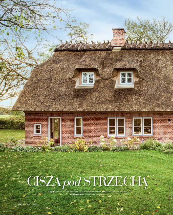 Ceglany wiejski dom z wysokim dachem krytym strzechą, jasnymi oknami i zielonym ogrodem. Na stronie otwierającej reportaż widnieje tytuł „Cisza pod strzechą”.
