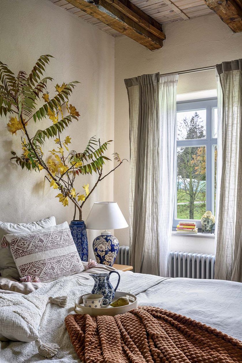 Przytulna sypialnia w mazurskim siedlisku z jasną pościelą, niebiesko-białą ceramiką, drewnianymi belkami i widokiem na ogród.