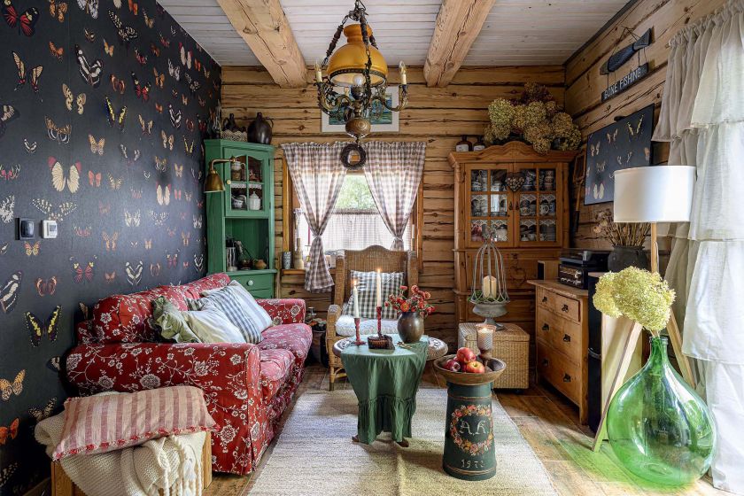 Przytulny salon w drewnianym domu z czerwoną sofą, tapetą w motyle, wiklinowymi meblami i rustykalnymi dekoracjami.