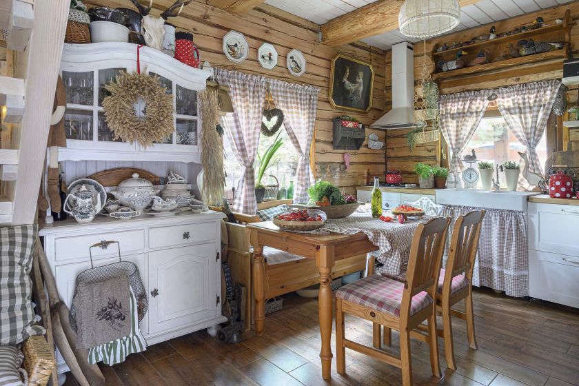 Rustykalna kuchnia w drewnianym domu z białymi meblami, stołem z litego drewna, zasłonami w kratę i dekoracjami w stylu country.
